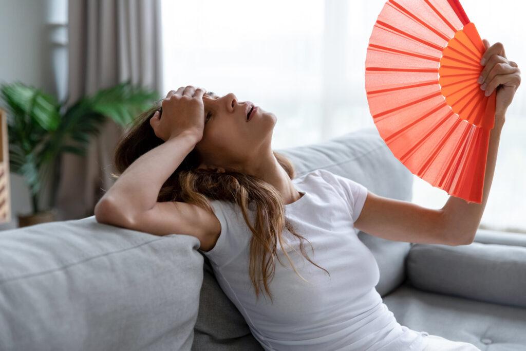 Erschöpfte Frau sitzt zuhause mit Fächer auf der Couch und leidet unter dem heißen Sommerwetter.