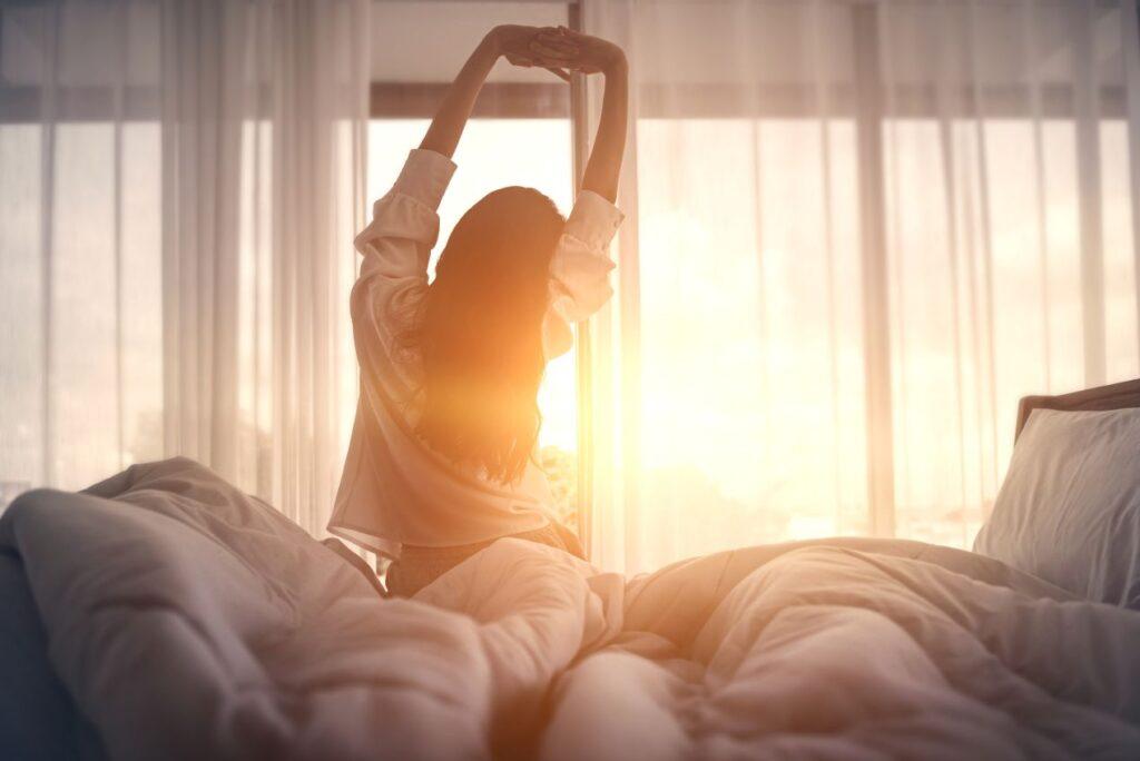 Frau streckt sich morgens in einem Bett mit Alpaka Bettdecke bei Sonnenaufgang